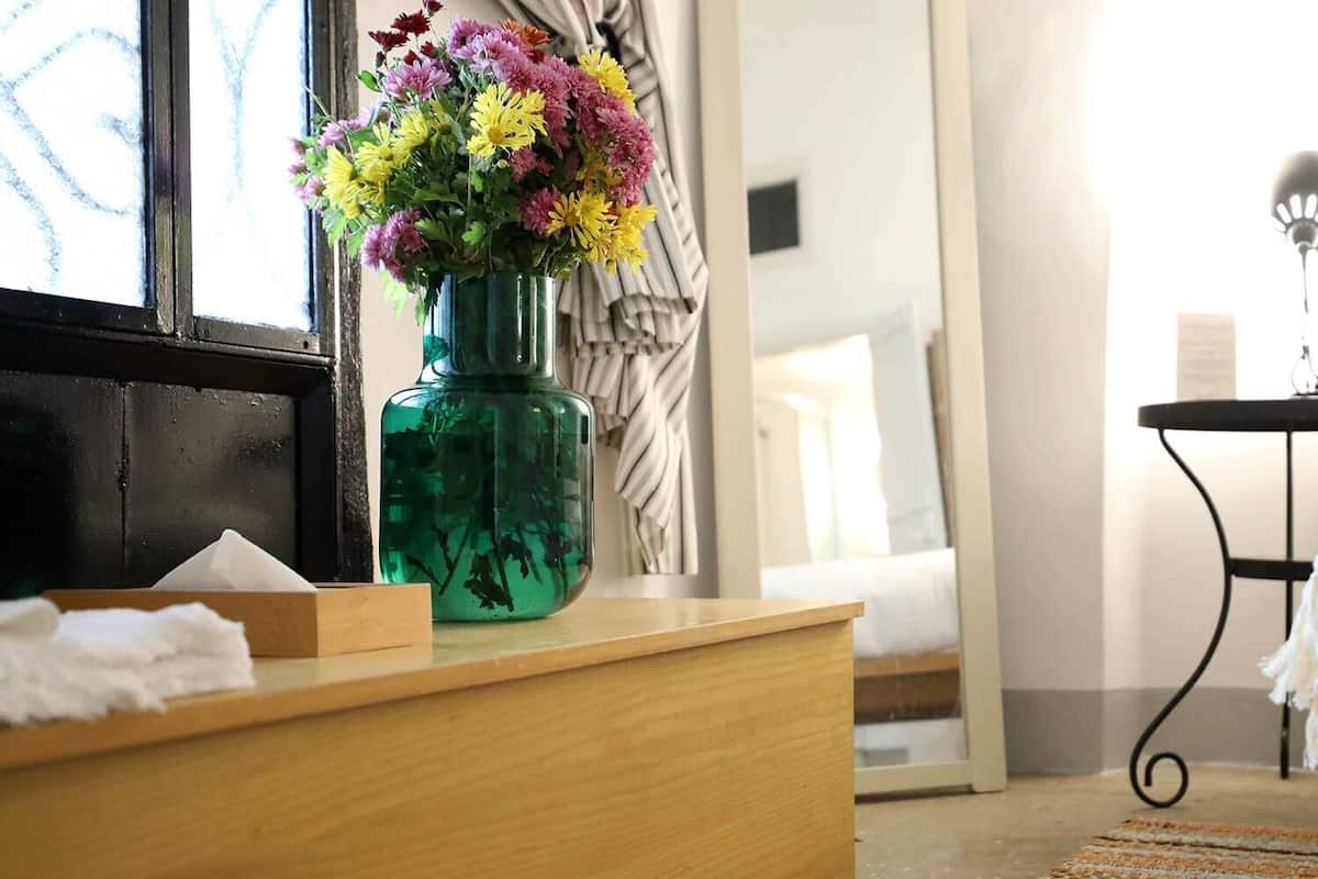 Hotellihuone, jossa on puinen työpöytä, vihreä maljakko täynnä kukkia, valkoisella peitolla varustettu sänky ja pieni pöytä, jossa on lamppu.