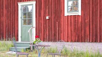 A red wooden building with a green door and a small window. There are two metal chairs and a table in front of the building.