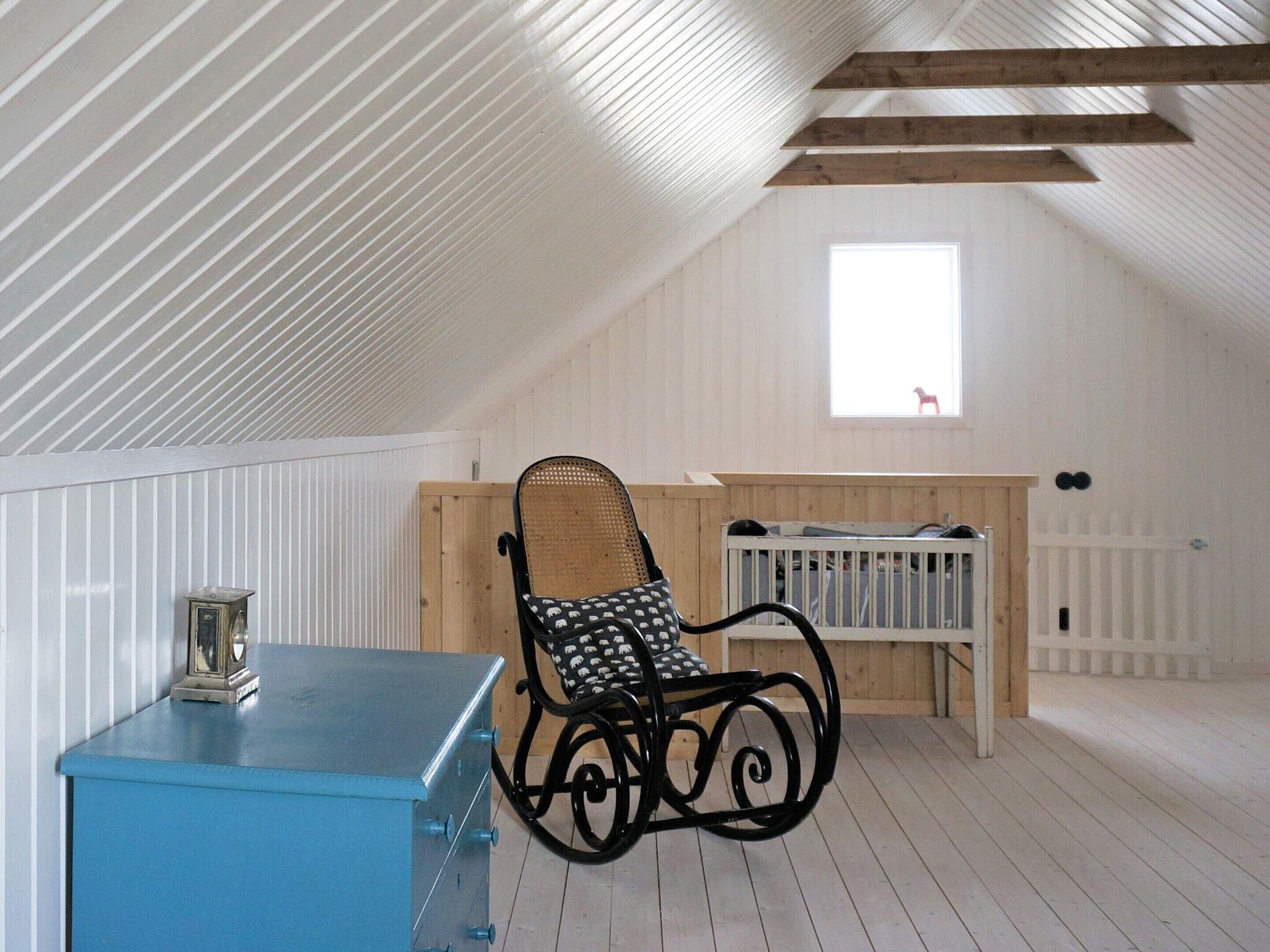 A room with a sloped ceiling, a blue chest of drawers, a rocking chair, a crib, and a window with a view of a red chair outside.
