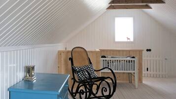 A room with a sloped ceiling, a blue chest of drawers, a rocking chair, a crib, and a window with a view of a red chair outside.