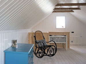 A room with a sloped ceiling, a blue chest of drawers, a rocking chair, a crib, and a window with a view of a red chair outside. - 4 Star Holiday Home in Nassjo (Nassjo)