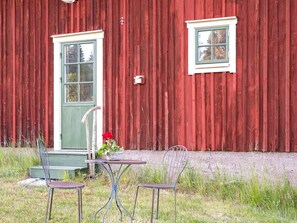 Un bâtiment en bois rouge, avec une porte verte et une petite fenêtre. On y trouve deux chaises en métal et une table devant le bâtiment.