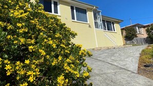 Une maison jaune à deux étages, avec le numéro « 22 » bien visible sur le côté, une allée en béton et un arbuste aux fleurs jaunes au premier plan.