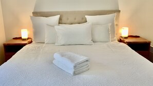 Un lit bien fait, avec une couette blanche, deux oreillers et une serviette pliée posée sur le lit. De chaque côté du lit, on trouve deux tables de chevet sur lesquelles sont posées des lampes.