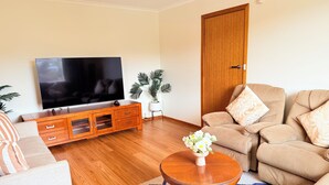 Un salon doté d’un téléviseur à écran plat, d’un meuble en bois, d’un canapé beige et d’une table basse ronde en bois sur laquelle est posé un vase de fleurs.