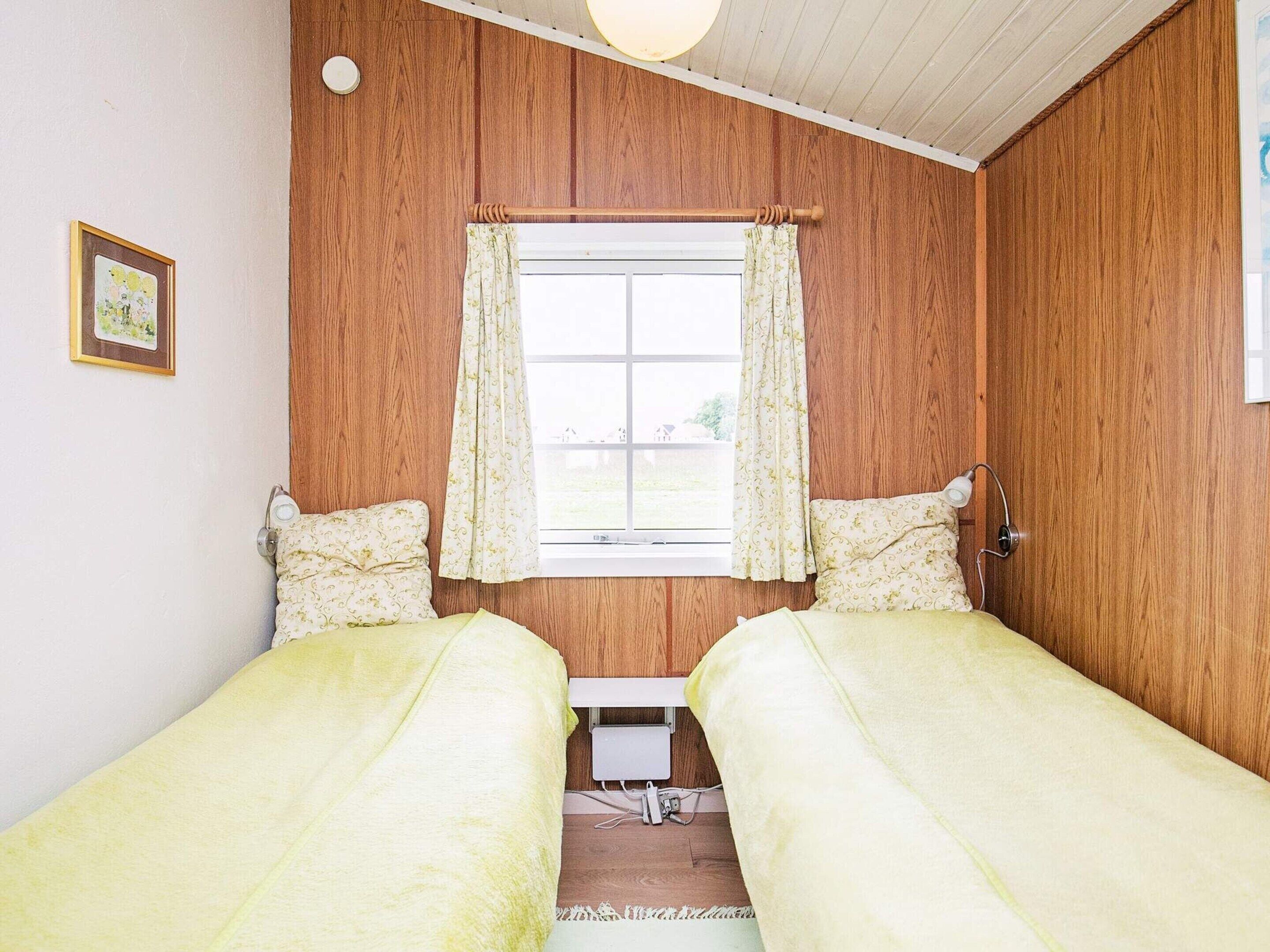 Una habitación con paredes de paneles de madera, una ventana con cortinas florales, dos camas con ropa de cama a juego, una mesita de noche con una lámpara y un cuadro en la pared.