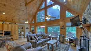 A spacious living room with a large wooden ceiling, a fireplace, and a television mounted on the wall. - Experience Lakeview Retreat, where pets are pampered by the shimmering waters. Unwind in a hot tub, enjoy WiFi, and  a full game room w/ pool table (Ellijay)