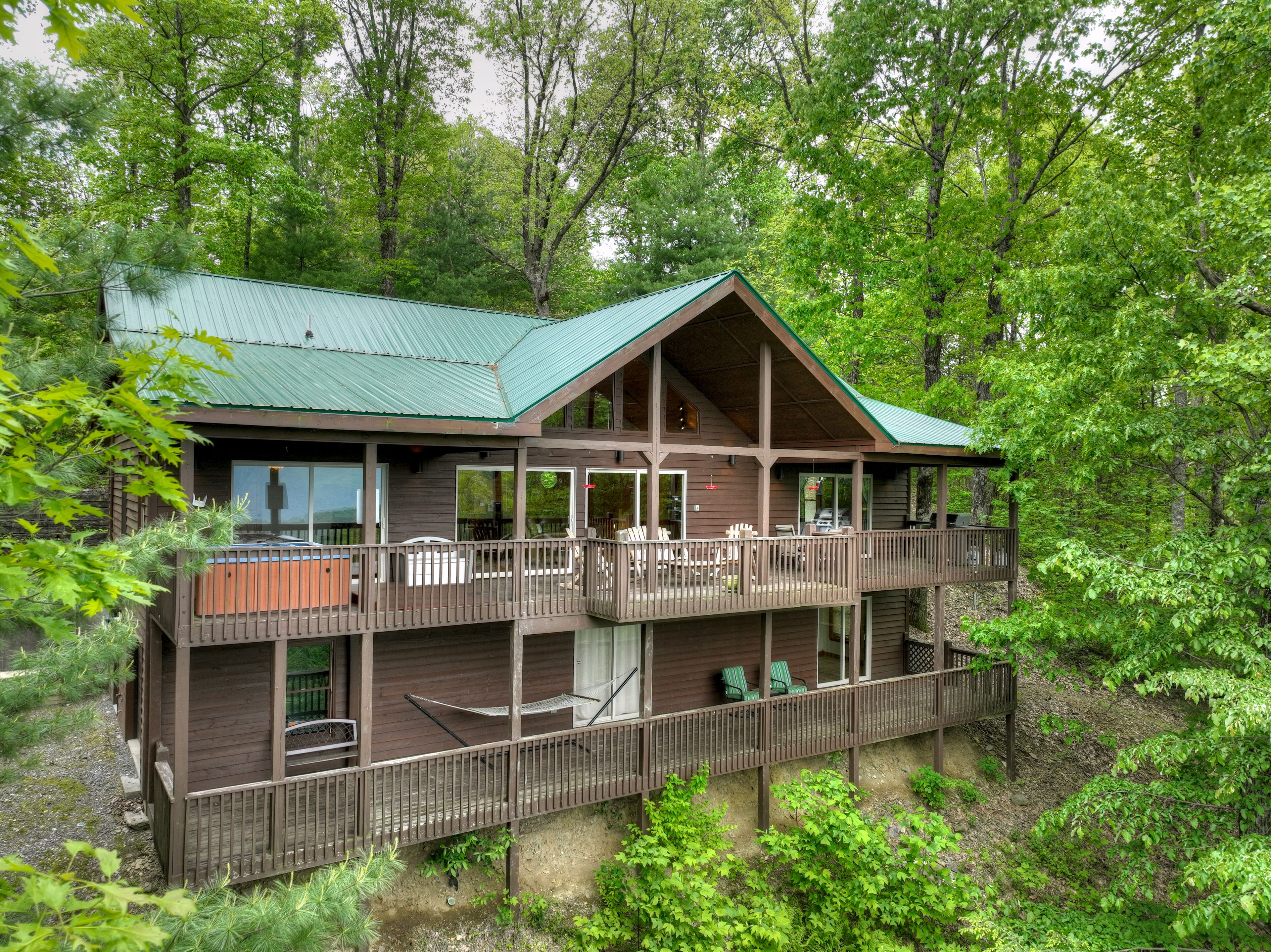 A two-story wooden cabin with a green roof, surrounded by dense forest.