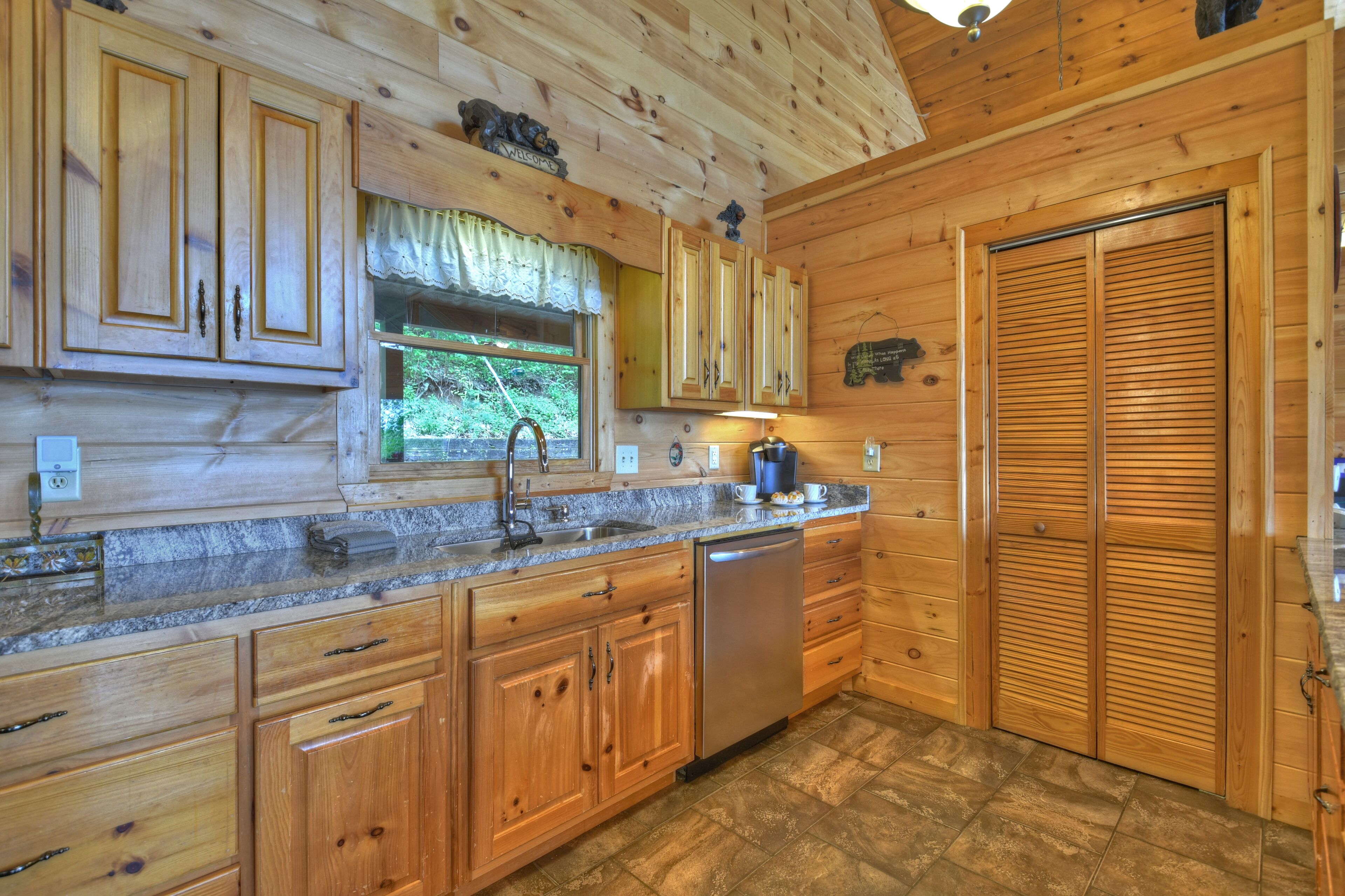 A wooden cabin kitchen with a granite countertop, wooden cabinets, a dishwasher, and a window with a curtain.
