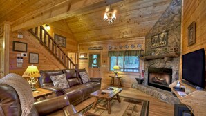 A cozy log cabin interior with a leather sofa, a fireplace, a television, and a staircase. - Misty Ridge - Where pets are pampered amidst stunning mountain panoramas, with outdoor fireplace, hot tub, firepit, pool table, and WiFi (Ellijay)