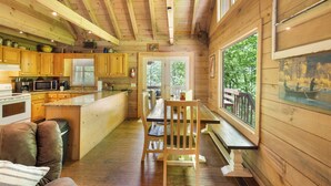 Interior de una cabaña con paredes de madera, una cocina con armarios de madera, una zona de comedor con un banco y un gran ventanal con vistas a la vegetación.