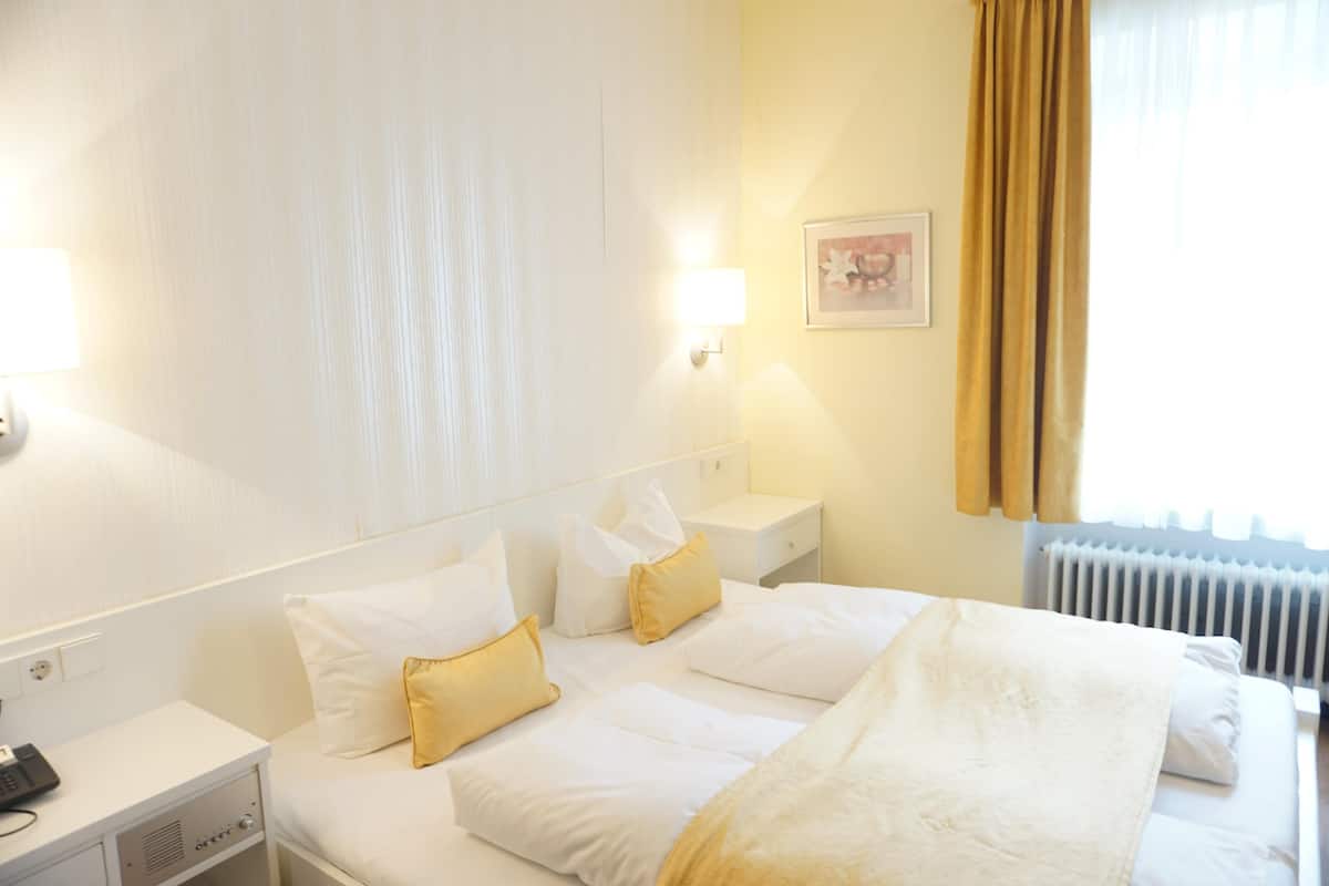 A hotel room with a bed, white linens, a bedside table with a lamp, a telephone, and a picture on the wall.