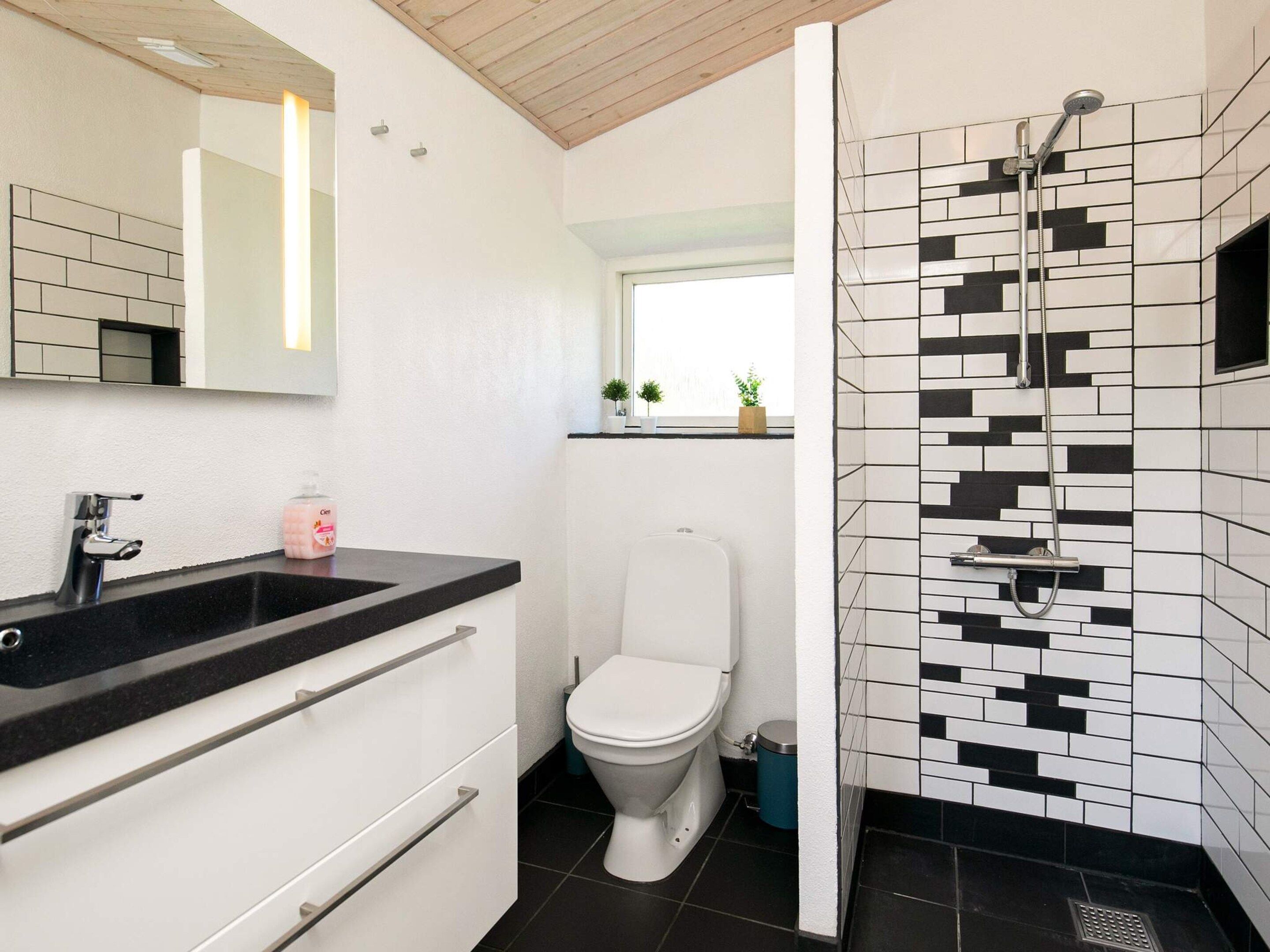 A modern bathroom with a black and white tiled shower, a white toilet, a black sink with a mirror above it, and a window above the toilet.