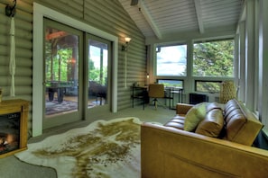 A cozy cabin interior with a leather sofa, a fireplace, and a large window overlooking greenery.