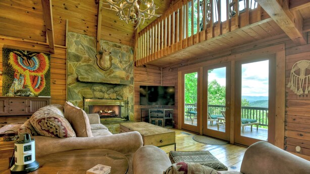 Interior acogedor de una cabaña con chimenea, paredes de madera y un balcón con vistas al exterior.