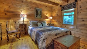 Un dormitorio de cabaña de madera con cama, una silla, una mesita de noche, una lámpara y una ventana.
