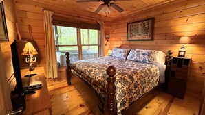 Un dormitorio de cabaña de madera con cama, ventilador de techo, ventana y mesita de noche con lámpara.