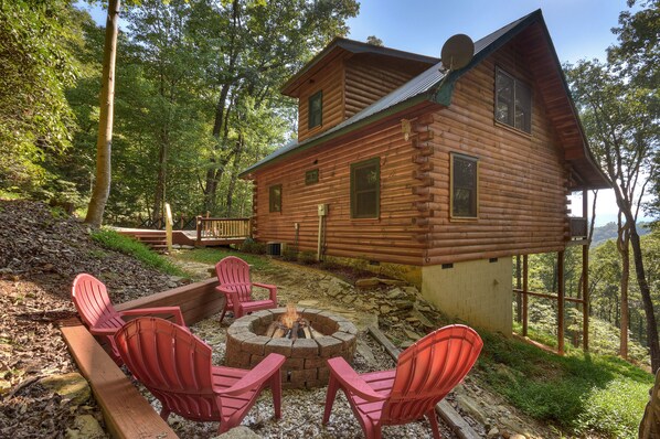 A log cabin with a fire pit and red chairs in the yard.