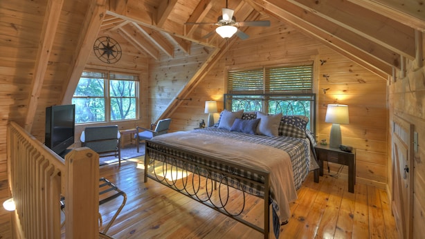 Um quarto de cabana aconchegante, com uma cama, uma cadeira, uma TV e uma janela com persianas.