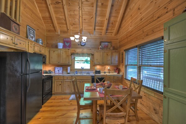 A wooden cabin kitchen with a dining area, wooden cabinets, a black refrigerator, and a window with blinds. - Indulge in Lofty Vista's stunning mountain panoramas. Relax in the hot tub, gather by the firepit, and enjoy hiking from your front door. WiFi (Blue Ridge)