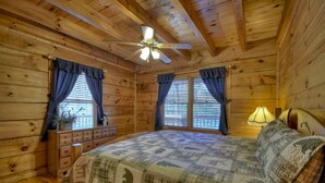 Un dormitorio de cabaña de madera con cama, cómoda, ventanas con cortinas, ventilador de techo y lámpara.