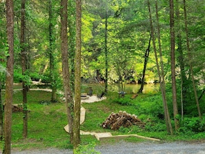 A forested campsite with a gravel path, a pile of logs, and a small clearing with a fire pit.