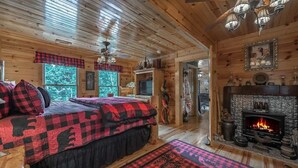 A cozy log cabin bedroom with a fireplace, a bed with red and black checkered bedding, and a view of the forest through large windows. - Experience the allure of Roost on the Ridge a pet-friendly sanctuary with hot tub, firepit, ping pong, spacious screen porch, and WiFi (Ellijay)