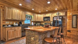 A wooden cabin-style kitchen with stone countertops, wooden cabinets, a central island with bar stools, a refrigerator, and a microwave.