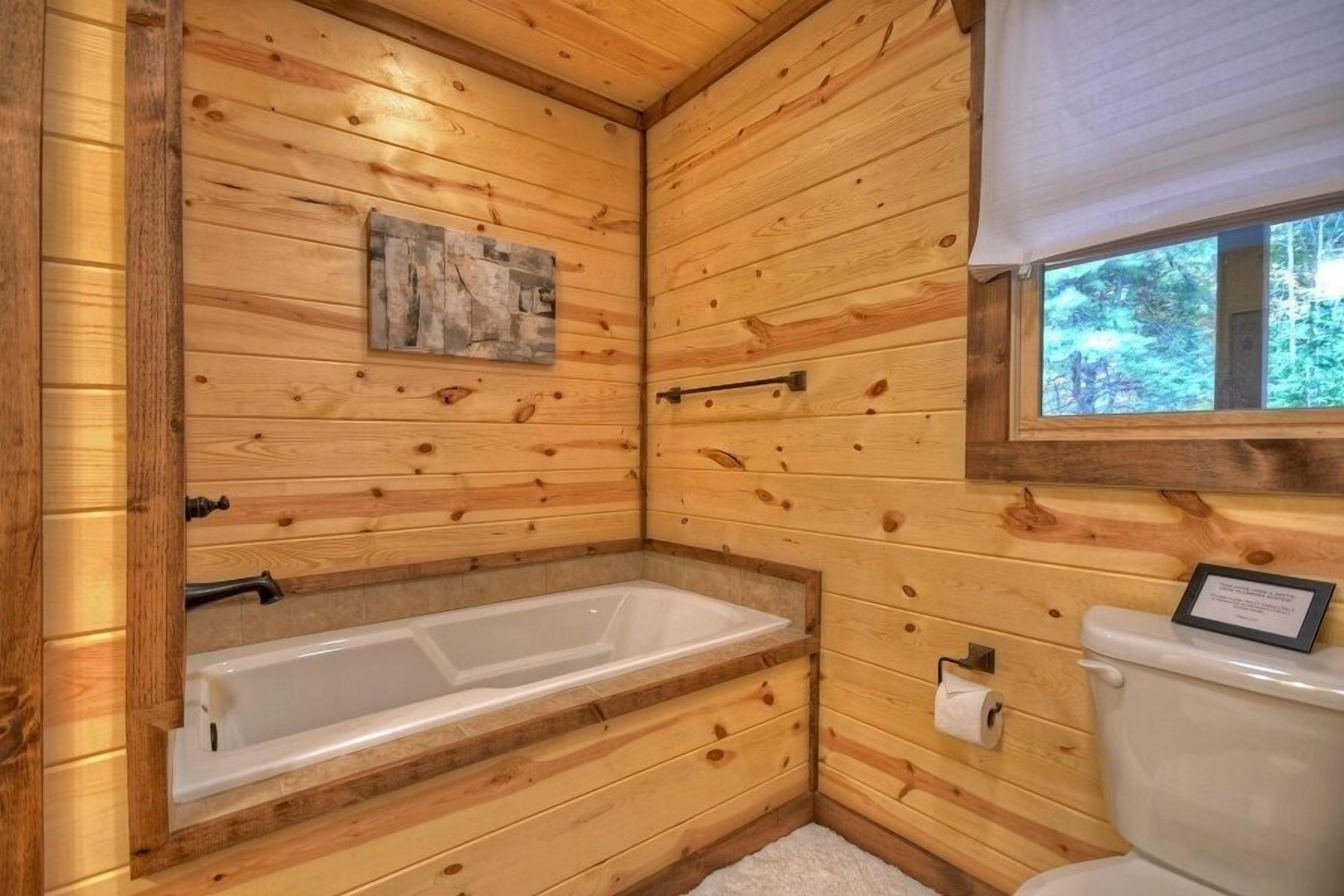 Une salle de bain rustique, avec des murs en bois, comprenant une baignoire, des toilettes et une fenêtre donnant sur la forêt.