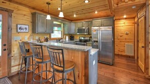 A wooden cabin interior with a kitchen featuring a central island, bar stools, and modern appliances. - Wakin on a Pretty Day: Luxurious pet-friendly retreat with stunning mountain views, hot tub, outdoor fireplace, pool table, WiFi, and downtown convenience (Blue Ridge)