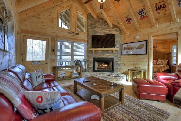 A cozy log cabin interior with a stone fireplace, leather furniture, and a television mounted above it.