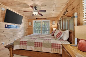 A wooden cabin room with a bed, a ceiling fan, a mounted TV, and a window with blinds.
