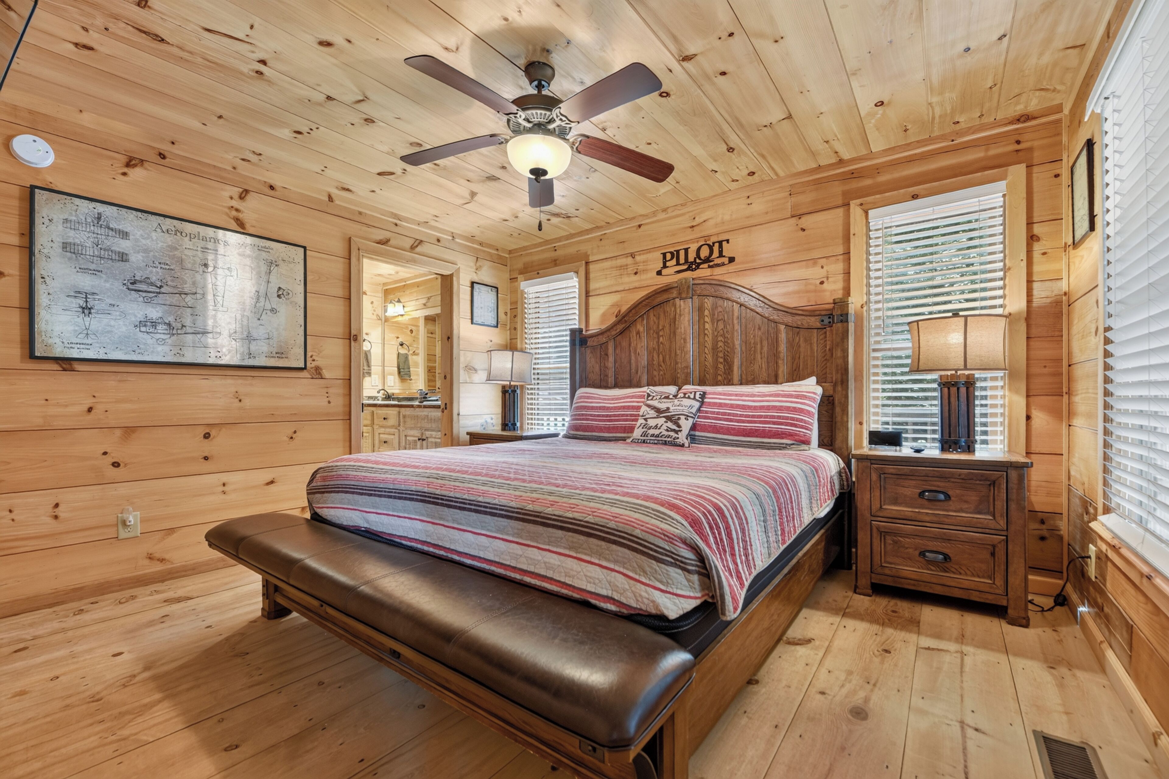 Une chambre dans une cabane en bois, comprenant un lit, un banc, un ventilateur de plafond et une carte d’aviation encadrée.