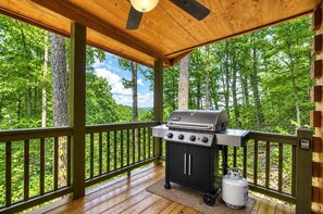 A covered outdoor area with a grill and propane tank, surrounded by a wooden deck and forest. - Up, Up and Away is pet friendly with a beautiful mountain view, hot tub, firepit, WiFi and fenced yard (Blue Ridge)