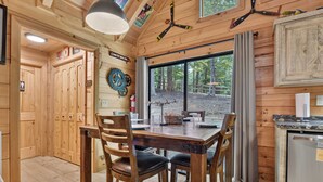 Een houten interieur met een eettafel, stoelen, een klok en een raam met uitzicht op bomen.