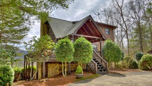 A log cabin with a steeply pitched roof, a wooden deck, and a staircase leading to a second-story balcony. - Ascend to serenity at Higher Ground! This pet-friendly haven offers majestic mountain views, a hot tub, firepit, WiFi, and unparalleled privacy. WiFi (Blue Ridge)
