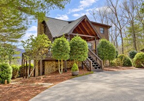 A log cabin with a stone chimney, a wooden staircase, and a paved driveway.