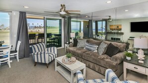A spacious living room with a large window, a ceiling fan, a sofa, a striped armchair, and a coffee table. - F14 Upper Cottage, Amazing oceanviews/Beachfront Luxury, Triple queen bunk, North Coast Village (Oceanside)