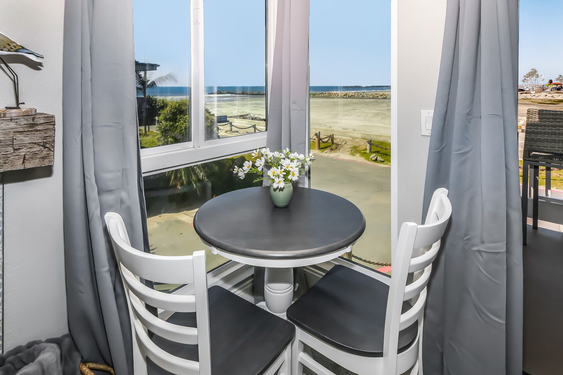 A corner room with a view of the beach, a small table with a vase of flowers, and white chairs.
