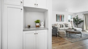 A modern living room with a grey sofa, a coffee table, and a rug. There is a white cabinet with a plant and a decorative item on top. - Remodeled /4 Car Parking/Clean/Patio w/ Firepit (Newport Beach)