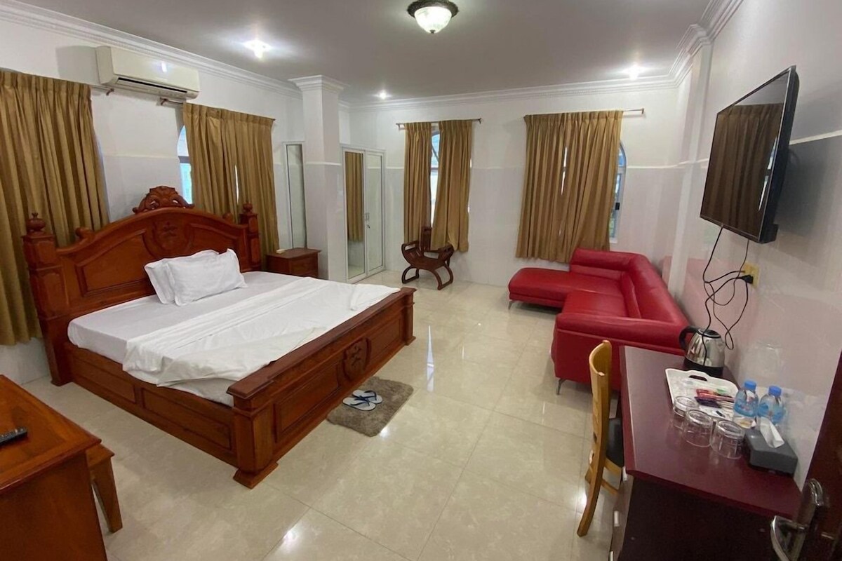 A hotel room with a wooden bed, a red sofa, a desk with a chair, a television, and an air conditioning unit.
