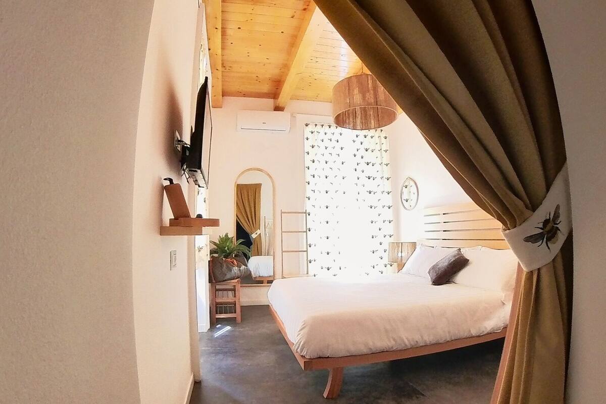 Quarto com cama de madeira, mesinha de cabeceira, planta em vaso e janela com cortinas.