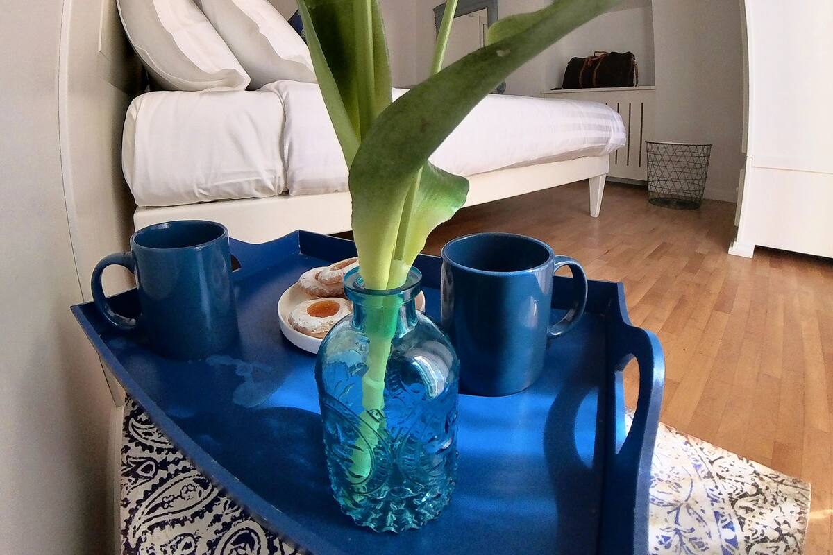 Quarto com cama, mesa de cabeceira com bandeja contendo um vaso e duas canecas, e janela com cortinas.