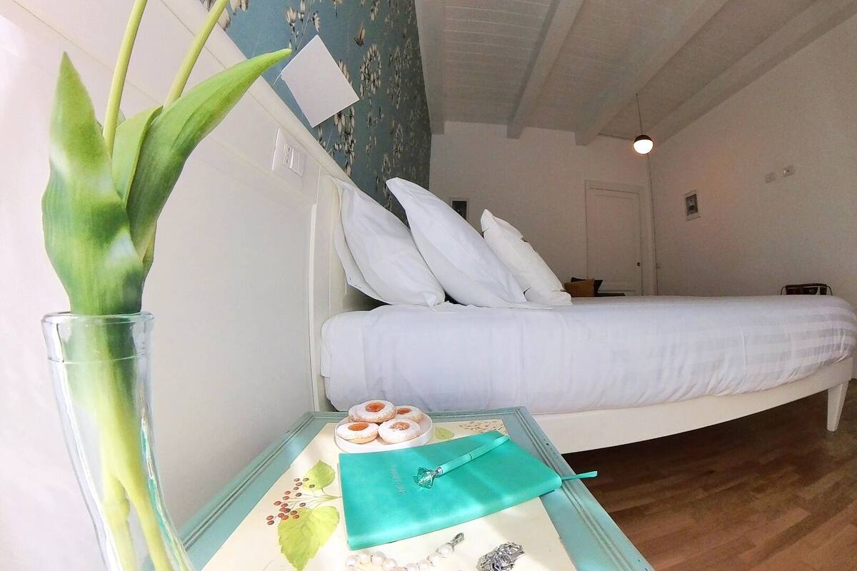 Quarto com cama, travesseiros brancos, mesa de cabeceira com um vaso de planta verde e bandeja com doces.