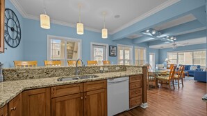 A spacious kitchen with granite countertops, wooden cabinets, and a dishwasher. There are multiple windows with blinds, a dining area with a table and chairs, and a blue sofa in the living room. - JUST UPDATED DEC '25!  4 br 4 ba ocean view villa- 16 steps to beach- pool club-directly across from beach access<br>Golf cart friendly. community pickle ball, tennis and basketball (Ocean Isle Beach)