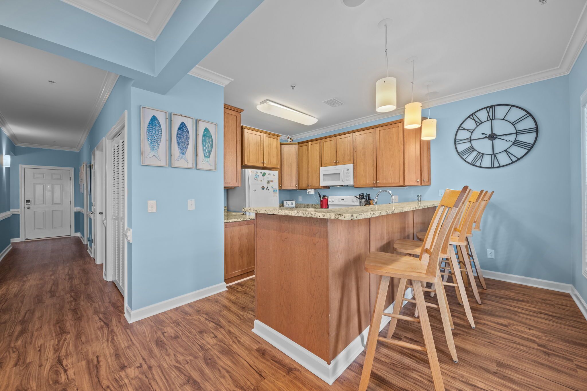 A kitchen with wooden cabinets, a central island with bar stools, a wall clock, and framed artwork on the wall.