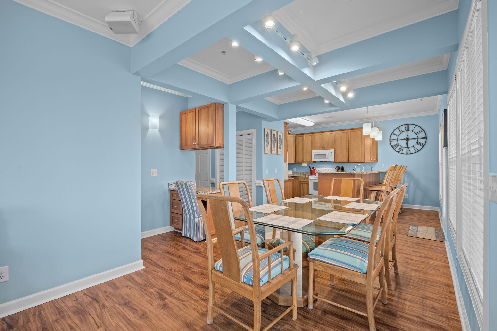 A dining area with a glass-top table, wooden chairs, and a kitchen in the background.