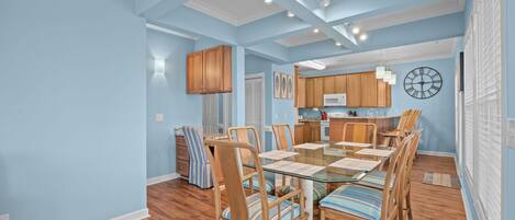 A dining area with a glass-top table, wooden chairs, and a kitchen in the background.