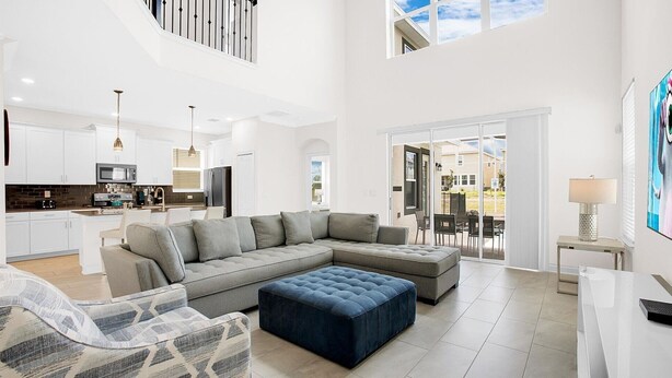 A spacious living area with a sectional sofa, a blue ottoman, and a dining area with a table and chairs. - Rent the Perfect 5 Bedroom House on Champions Gate Resort, Orlando House 6166 (Kissimmee)