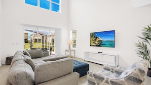 A spacious living room with a large sectional sofa, a flat-screen TV mounted on the wall, and a view of a tropical beach through large windows. - Rent the Perfect 5 Bedroom House on Champions Gate Resort, Orlando House 6166 (Kissimmee)